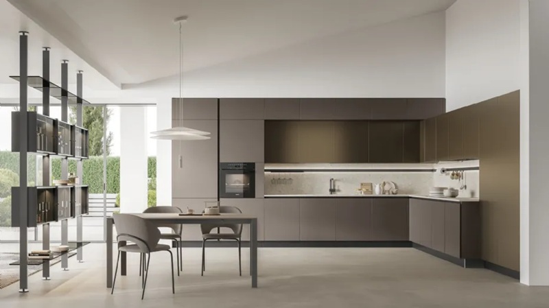 Cucina Stosa angolare con ante bronzo opache, piano chiaro, forno incassato, tavolo con sedie grigie e libreria divisoria