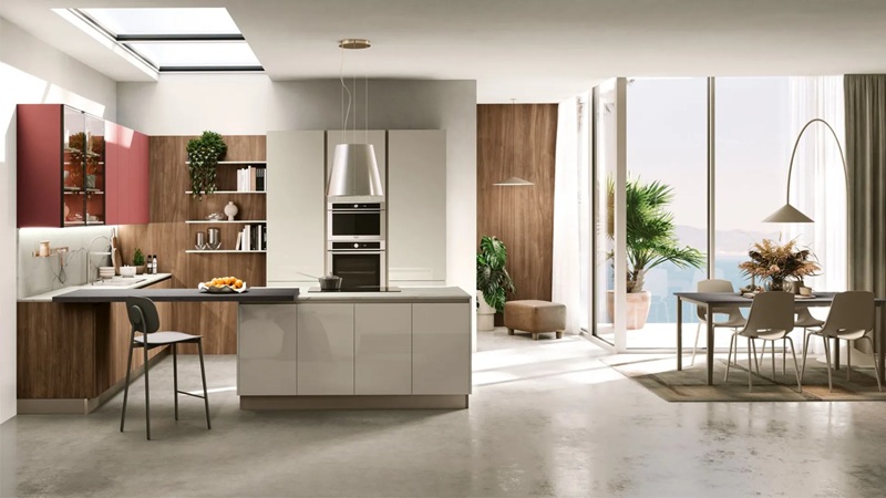 Cucina open space con isola grigio lucido, pensili rossi e boiserie in noce, accanto a tavolo con sedie beige e vetrate