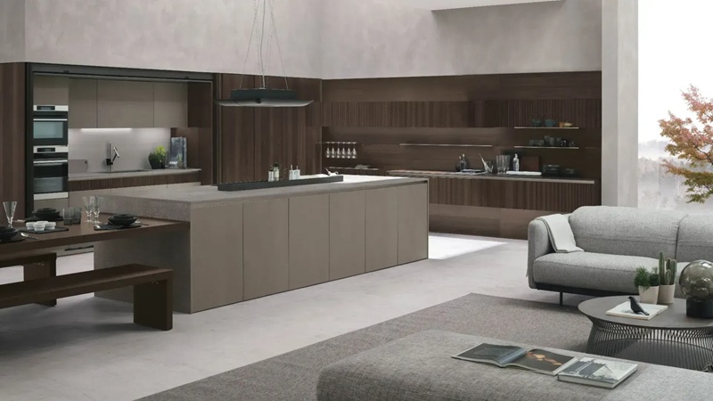 Cucina Stosa open space con isola color tortora, pareti in legno scuro, forni da incasso e divano grigio